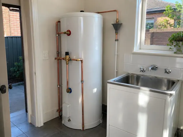 White hot water cylinder with copper pipework beside a laundry sink and window in Petersham